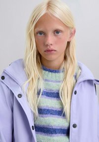 Chaqueta impermeable morada sobre un suéter a rayas multicolor con tonos verdes, azules y morados. Textura de tela suave y botones visibles.