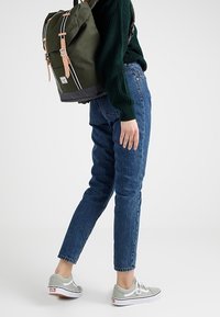 Grøn lærredsrygsæk med læderdetaljer, rullet top og hvide striber. Modellen har en mørk sweater, blå jeans og grå sneakers på.