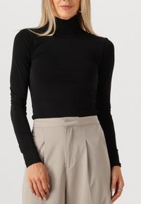 Zwart turtleneck shirt met lange mouwen van rekbare stof, gecombineerd met lichtbeige high-waisted broeken met plooien en een aangepaste pasvorm.