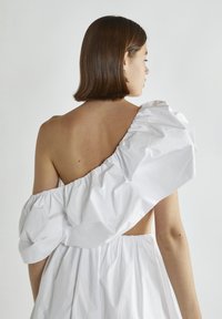 Hvid kjole med en oversized, voluminøs ærme; åben rygdesign. Stoffet ser tekstureret og let ud. Model set fra bagfra.