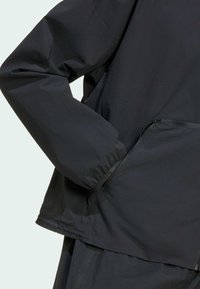 Crna jakna s teksturiranim završetkom, koja uključuje džep s patentnim zatvaračem i elastične rukave, dizajnirana za funkcionalnost i udobnost.