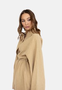 Beżowy, oversize płaszcz zapinany na pasek, z szerokimi rękawami i teksturowanym materiałem. Składany kołnierz podkreśla jego luźną strukturę.