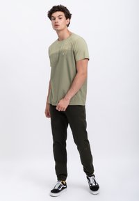 Olive green cotton t-shirt with short sleeves and subtle chest design. Paired with dark green tapered pants and black sneakers with white accents.