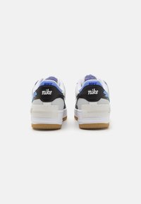 Bílé a černé Nike tenisky s modrými akcenty. Mají texturovaný podpatek, polstrovaný jazyk a gumovou podrážku. Viditelné logo "Nike".