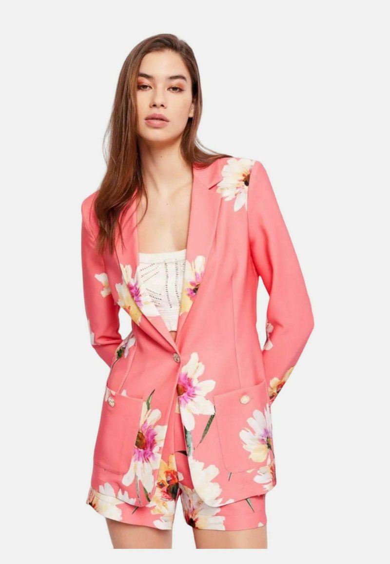 Blazer floreale rosa con due tasche frontali e pantaloni coordinati, caratterizzato da grandi fiori bianchi e accenti verdi su un tessuto liscio.