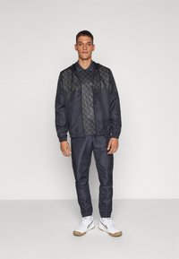 Chaqueta cortavientos navy con un panel en el pecho estampado, combinada con pantalones a juego. Ambos tienen una textura suave y dobladillos elásticos. Zapatillas deportivas blancas.