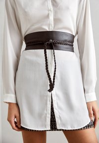 Witte blousejurk met een split, gecombineerd met een brede donkerbruine gevlochten leren riem die om de taille is geknoopt en een patroonrok eronder.