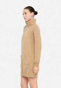 Abito smanicato beige a collo alto in maglia con texture a coste, tasche laterali e una vestibilità morbida, che arriva a metà coscia. Finitura in tessuto liscia.
