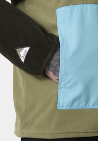 Grüne Fleecejacke mit einer hellblauen Tasche, schwarzen Ärmeln mit einem weißen Logo und strukturiertem Stoffdetail.