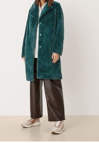 Turkusowy płaszcz z futra syntetycznego z dużym kołnierzem i zapięciem na guziki, skomponowany z brązowymi spodniami z ekoskóry i beżowymi sneakersami z białymi akcentami.