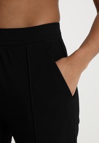 Svarta byxor med stretchmidja, sidofickor och veck vid midjan för extra detalj. Mjuk och slät tygkvalitet.