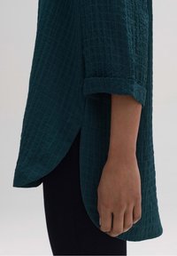 Długosleeve tunika w ciemnym teal, z teksturowanym wzorem w kratkę. Bocznie rozcięcia i podwinięte mankiety podkreślają luźny fason.