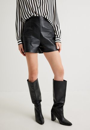 Pantaloni corti neri in pelle a vita alta con superficie liscia, abbinati a stivali neri al ginocchio. Camicia a maniche lunghe a righe bianche e nere visibile.