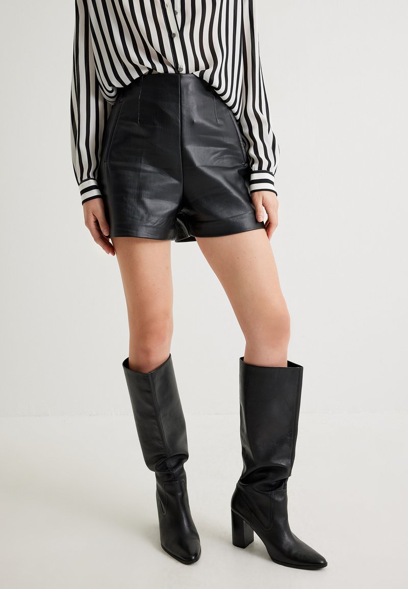 Pantalones cortos de cuero negro de cintura alta con textura suave, combinados con botas negras hasta la rodilla. Camisa de manga larga a rayas en blanco y negro visible.