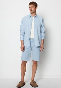 Lichtblauw linnen overhemd met knopen, wit t-shirt, lichtblauwe shorts, bruine instappers, staand tegen een effen achtergrond.