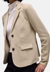 Blazer beige texturé avec un motif tissé, doté d'un col à revers, de deux boutons et d'une poche avant unique. Porté sur un col roulé blanc.