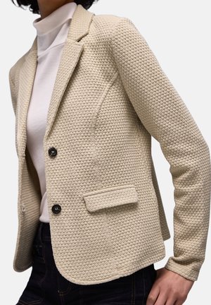 Beige strukturierter Blazer mit gewebtem Muster, ausgestattet mit einem Reverskragen, zwei Knöpfen und einer einzelnen Vorderseite Tasche. Wird über einem weißen Rollkragenpullover getragen.