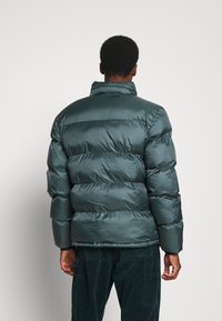 Persona che indossa un giubbotto trapuntato di un verde teal scuro e pantaloni di corduroy scuri, mostrata di spalle su uno sfondo chiaro e uniforme.