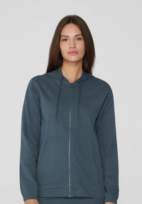 Sweatshirt à capuche zippé en bleu foncé. Il dispose d'une poche avant, de poignets côtelés et d'une texture douce et lisse. Cordons au niveau de la capuche.