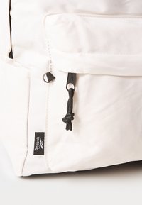 Weißer Canvas-Rucksack mit einem schwarzen Reißverschluss, verstellbarem schwarzen Kordelzug und einem dezenten Logopatch. Strukturierte Materialien mit einem klaren Design.