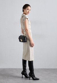 Beiges, figurbetontes Kleid mit kurzen Ärmeln, kombiniert mit schwarzen kniehohen Stiefeln mit Stiletto-Absätzen. Eine kleine schwarze, gesteppte Tasche mit einer goldenen Kette ergänzt das Outfit.