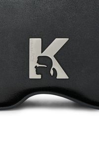 Črn teksturiran material z zlatim kovinskim logotipom v obliki črke K, ki vključuje silhuetno obliko in ime blagovne znamke spodaj.