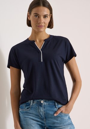 Femme aux cheveux courts et bruns portant un haut bleu marine à manches courtes avec bordure blanche et un jean bleu, debout avec une main dans la poche.