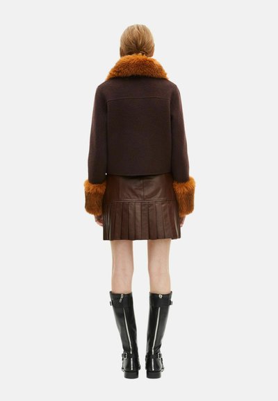 Chaqueta marrón recortada con cuello de piel sintética naranja, combinada con una falda de cuero marrón con pliegues y botas de cuero negras hasta la rodilla.