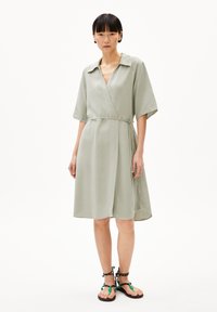 Light green wrap dress with short sleeves, tie waist detail, flowing fabric, and a subtle sheen. Paired with black and green strappy sandals.