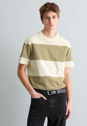 Young man wearing a green and white striped short-sleeve shirt, black jeans with a black belt, silver bracelet and chain necklace, standing.