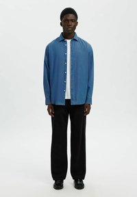 Ljust blå skjorta med krage, lagd över en vit skjorta. Bärs med svarta byxor med breda ben och svarta skor, stående mot en vit bakgrund.