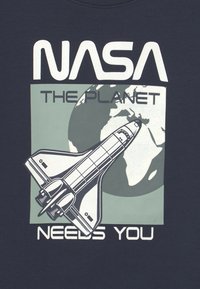 Tričko s potiskem inspirovaným NASA, na tmavě modrém pozadí s designem raketoplánu v bílé barvě, s nápisem "PLANETA TĚ POTŘEBUJE" a ilustrací země.