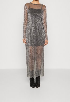 Kvinde iført en lang, gennemsigtig sølvglitrende kjole over en sort underkjole med sorte ankelstøvler, stående foran en ensfarvet baggrund.