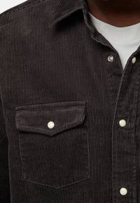 Zbliżenie ciemno-brązowej koszuli z kordurowej tkaniny z klapką na kieszeń i białymi zatrzaskami, noszonej na białej koszulce z okrągłym dekoltem.