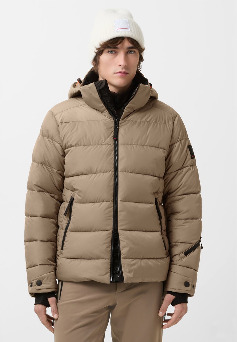 Beige puffer jacket with a hood, featuring horizontal quilting, black zippers, and side pockets. Worn with a white knit beanie.