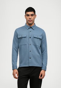 Modrá košile s dlouhým rukávem a zapínacím límcem, dvěma náprsními kapsami a tmavými knoflíky. Vyrobena z texturované látky s přiléhavým střihem.