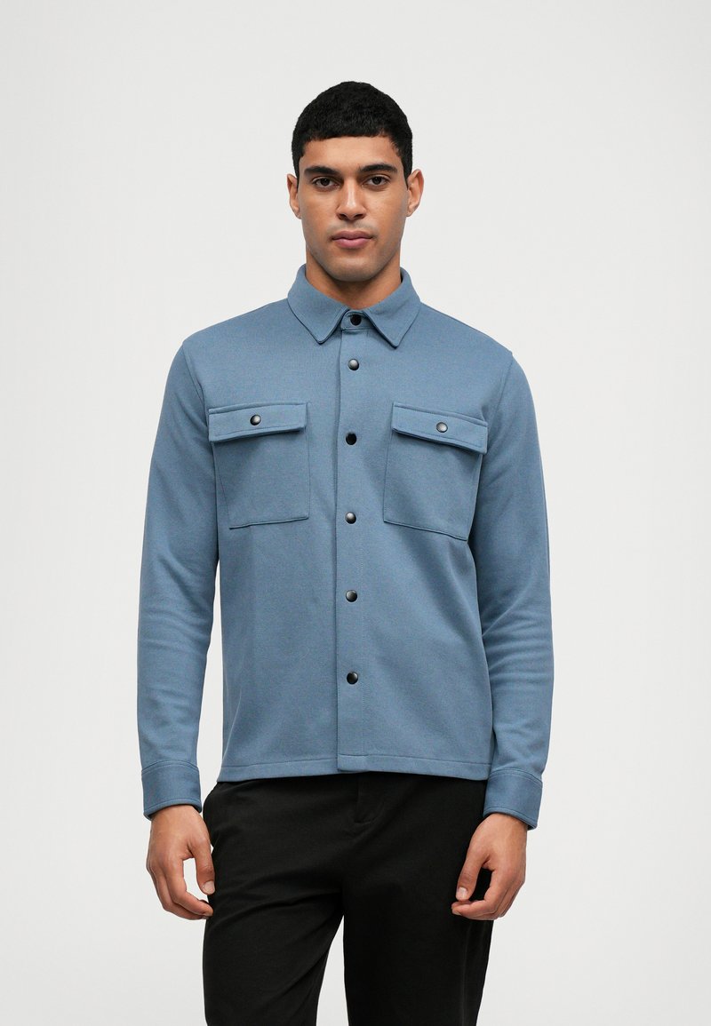 Modrá košile s dlouhým rukávem a zapínacím límcem, dvěma náprsními kapsami a tmavými knoflíky. Vyrobena z texturované látky s přiléhavým střihem.