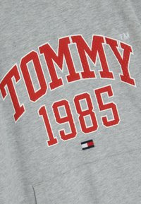 Felpa grigia con grande scritta rossa "TOMMY" e "1985." Include un piccolo logo ricamato a bandiera nei colori blu, rosso e bianco. Tessuto morbido.