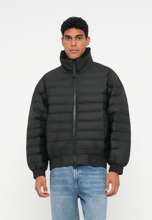 Man i svart vadderad jacka med hög krage och blå jeans, stående mot en enfärgad vit bakgrund.