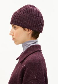 Geribbelde gebreide beanie in dieppaars met kleurvlekken. Zachte stof, ronde bovenkant, gedragen met een hooggesloten grijze top. Profielweergave.