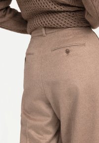 Närbild på beige skräddarsydda ullbyxor med knäppning på bakfickorna, bära tillsammans med en strukturerad brun stickad tröja.