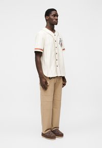 Μπλούζα μπέιζμπολ με κοντά μανίκια σε κρεμ απόχρωση με μαύρα κουμπιά και κόκκινες ρίγες, σε συνδυασμό με μπεζ παντελόνια cargo και καφέ παπούτσια με σχέδια.