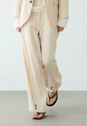 Person iført beige bukser med brede ben med knapdetaljer ved kanterne, beige blazer og sorte thong-sandaler, der går på gråt gulv.