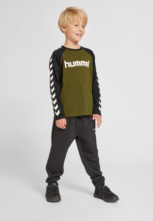 Olivegrünes Langarm-T-Shirt mit schwarzen Ärmeln, mit weißem "hummel"-Logo und Chevron-Muster. Schwarze Hose und schwarze Turnschuhe.