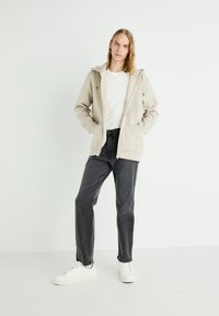 Sudadera con cremallera de color beige, con capucha, bolsillos laterales y detalles de cremallera negra, combinada con jeans gris oscuro y zapatillas blancas.