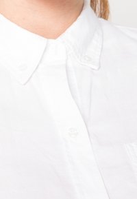 Gros plan sur une chemise blanche à col boutonné portée par une personne à la peau claire et aux cheveux bruns visibles.