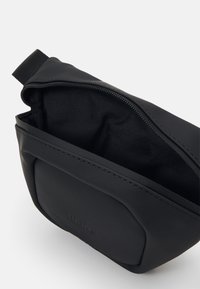 Crna vodootporna torba napravljena od glatkog materijala s zatvaračem i teksturiranim nylon unutrašnjim dijelom. Ima mali brendirani detalj sprijeda.