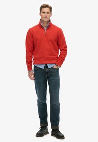 Suéter rojo con cuello con cremallera sobre una camisa de botones azul claro, combinado con jeans azul oscuro y zapatos negros con cordones.