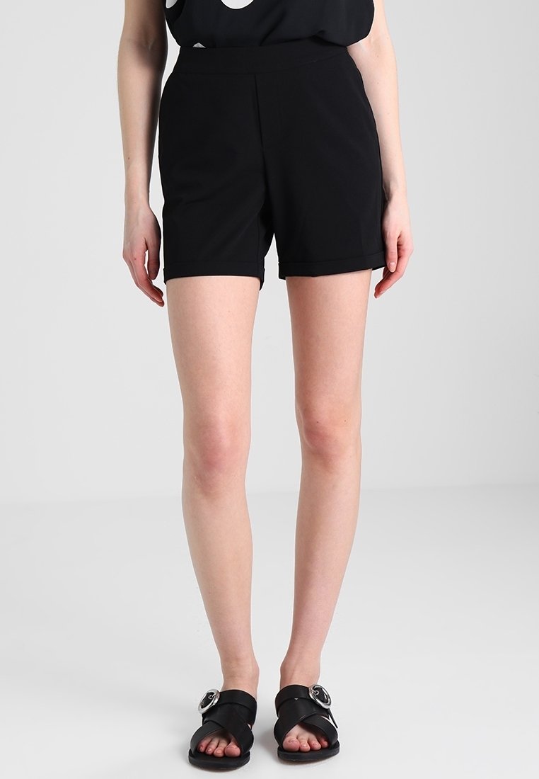 Shorts noirs à texture lisse, longueur mi-cuisse, avec une ceinture ajustée. Portés avec des sandales plates noires ornées de ferrures décoratives.