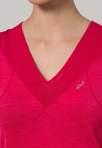 Jasnoróżowa koszulka sportowa z dekoltem w kształcie litery V, z warstwowym wyglądem i srebrnym logo po lewej stronie. Wykonana z miękkiego materiału.